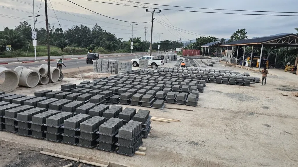 Concrete blocks stacked on pallets across the roadside production yard, with concrete pipes and open-sided sheds